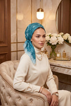 Load image into Gallery viewer, A woman wearing a teal blue patterned headscarf and a cream turtleneck sweater sits in a tufted beige armchair in an elegant room with marble walls, a vanity table, and a vase of white flowers.