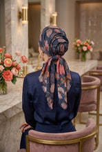 Load image into Gallery viewer, A woman seated with her back to the camera wearing a navy blue satin top and a dark floral headscarf with pink and rust accents, in a modern marble bathroom or dressing area with pink velvet chairs.
