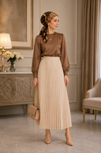 Load image into Gallery viewer, A woman stands in an elegant room wearing a brown satin long-sleeve blouse, a pleated cream midi skirt, and a leopard-print headscarf, holding a beige handbag.
