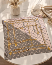 Load image into Gallery viewer, A square silk scarf with a luxury monogram-inspired design in pink, gray, and gold, styled on a round white marble table with a vase and dried grass in the background.
