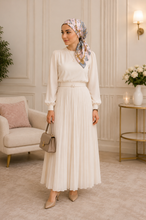 Load image into Gallery viewer, A woman stands in a softly lit interior wearing a cream long-sleeve blouse and matching pleated maxi skirt with a thin waist belt, paired with a floral headscarf and beige pointed heels. She holds a taupe handbag.

