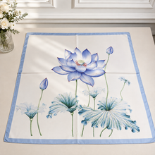 Load image into Gallery viewer, A square white tablecloth with a light blue border and a watercolor-style lotus floral print in shades of blue, displayed on a light beige tabletop.
