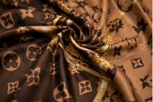 Load image into Gallery viewer, A collage showing a woman modeling a brown and gold monogram scarf/headscarf in multiple views, including side profiles, a close-up of the fabric pattern, and a full-body pose in an indoor room. The scarf features an ornate monogram print in gold on a dark brown background.
