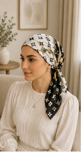 Load image into Gallery viewer, A woman wearing a black and white monogram patterned silk headscarf with gold chain accents, styled in a warm, neutral-toned indoor setting.
