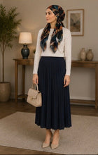Load image into Gallery viewer, A woman stands in a styled indoor room wearing a cream long-sleeve top, a navy pleated midi skirt, and a multicolor patterned hijab. She holds a beige handbag and wears beige heels.
